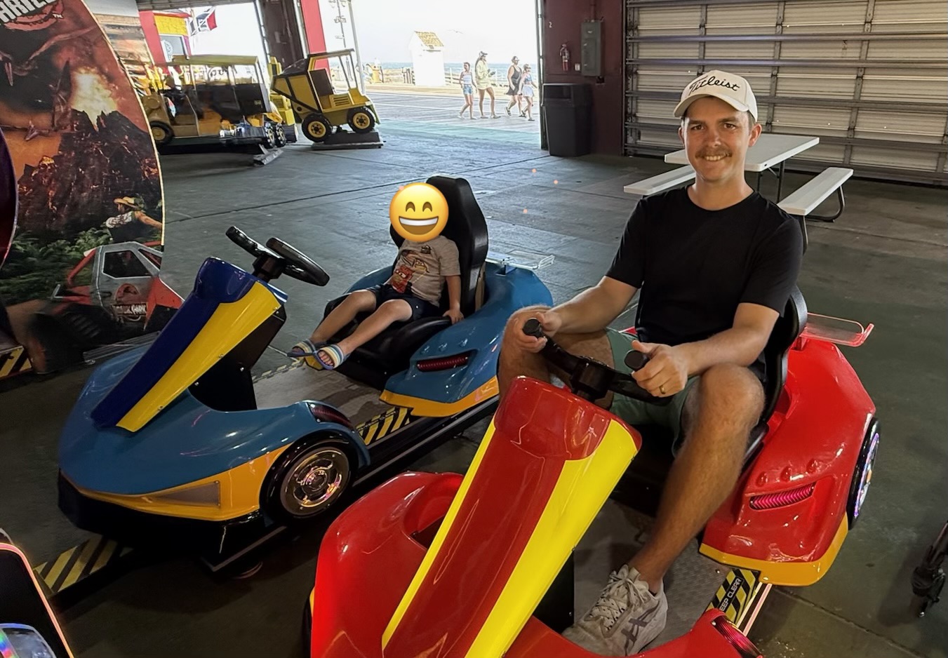Jamey and his son seated side by side in small ride-on karts at an indoor arcade.
